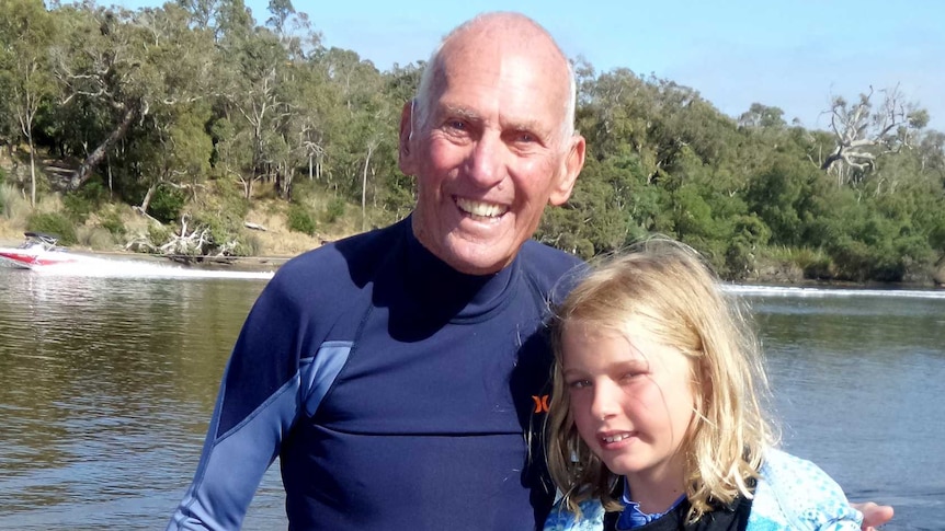 An elderly man wearing swimming gear with his arm around a young girl in front of a river.