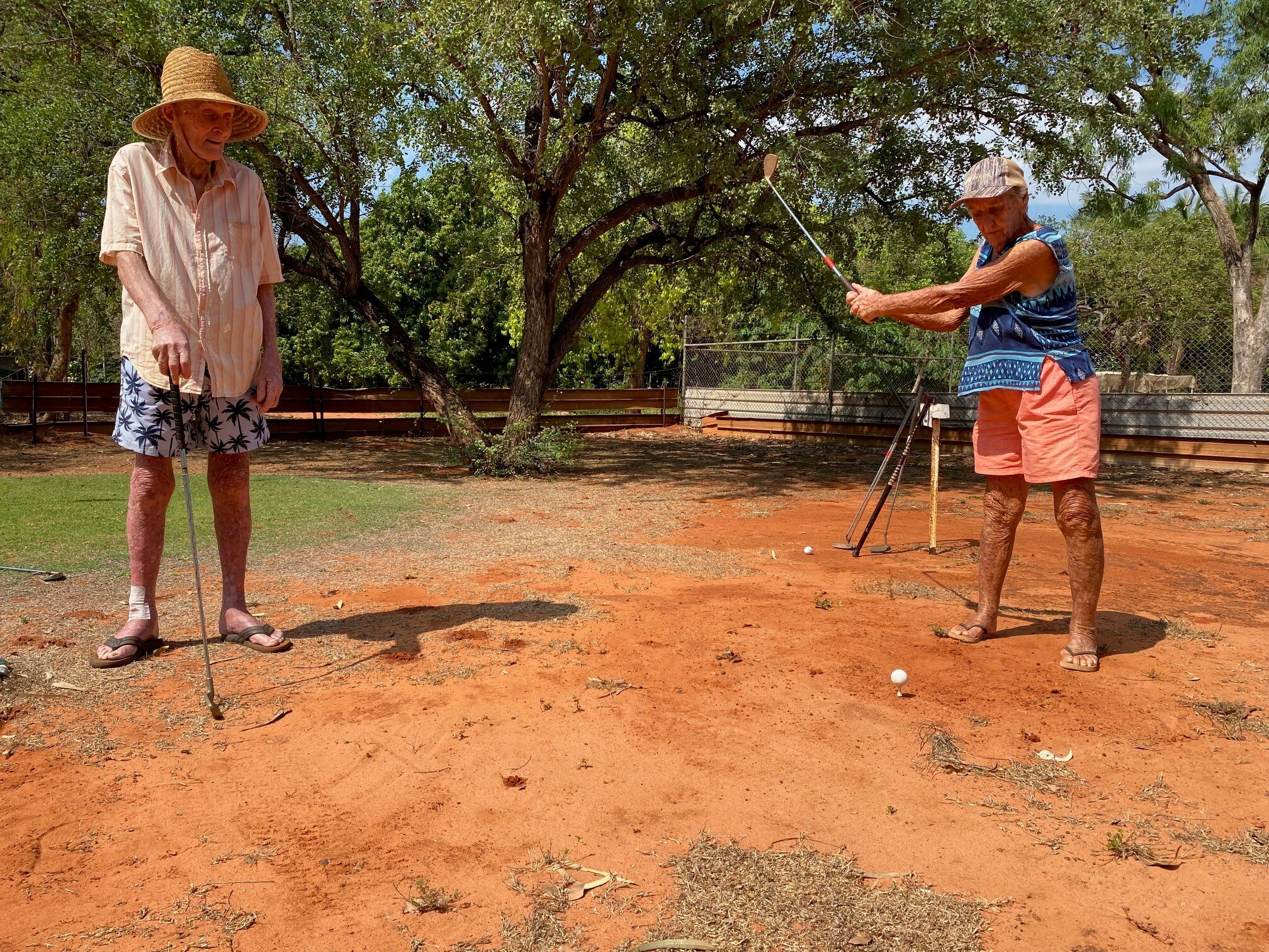 A woman swings her golf club at a ball on red soil while a man in a hat watches