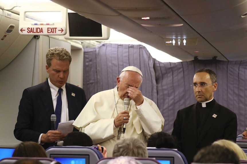 Pope Francis has one hand on his face and is flanked by father Mauricio Rueda and Greg Burke, spokesman of the Vatican.