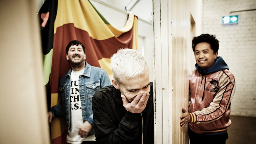 The Avalanches situated around doorway, Robbie Chater laughs into his hand in foreground, Tony and James flank him in background