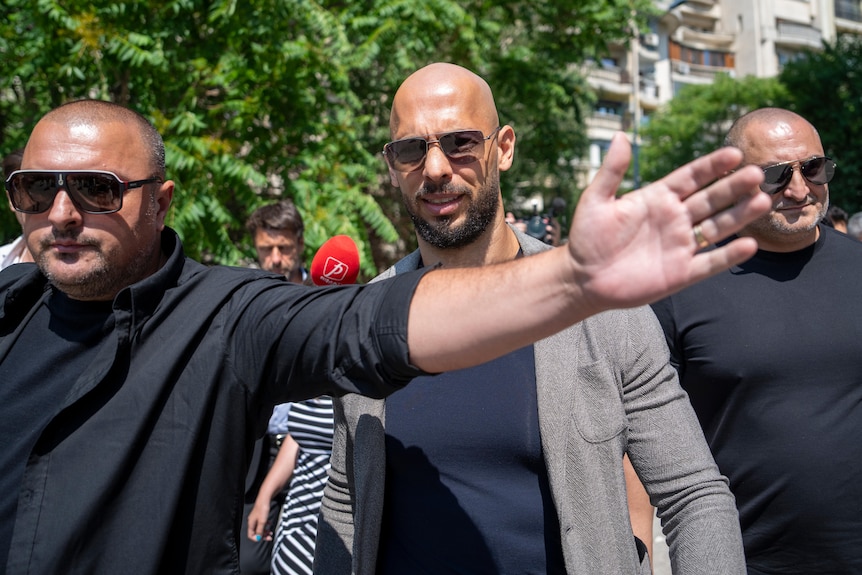 Andrew Tate is pictured, two bald men with sunglasses walk besides him while one raises his arm seemingly to protect Tate.