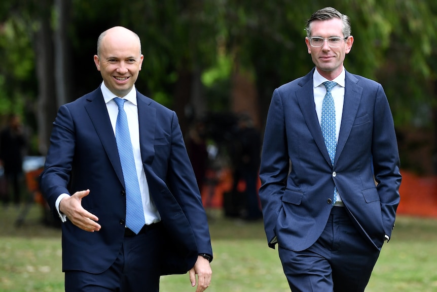 two men walking in a park, the one on the left has his right hand out, as if he's about to shake hands with someone