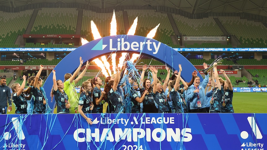 A women's soccer team wearing dark blue celebrates winning a trophy in front of a big blue sign saying "champions"
