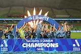 A women's soccer team wearing dark blue celebrates winning a trophy in front of a big blue sign saying "champions"
