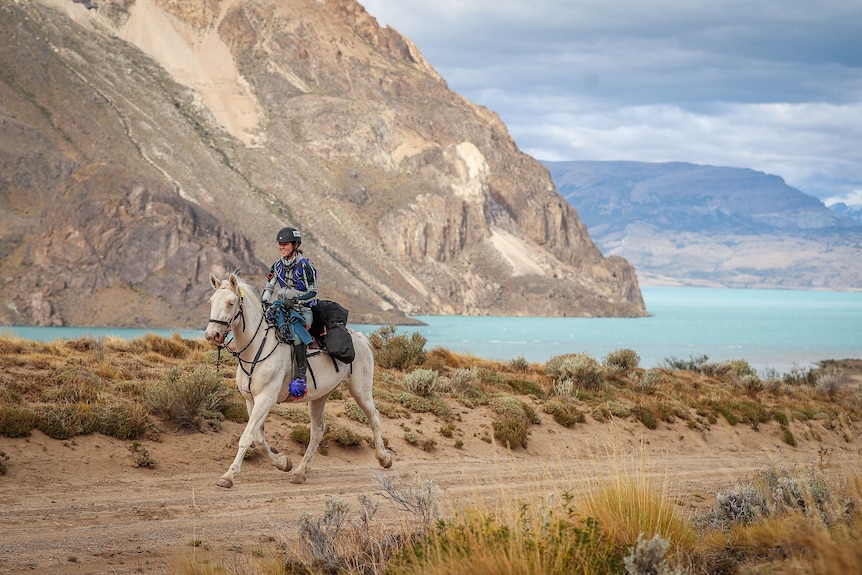 A rider on a white horse passes a blue lake and mountains.