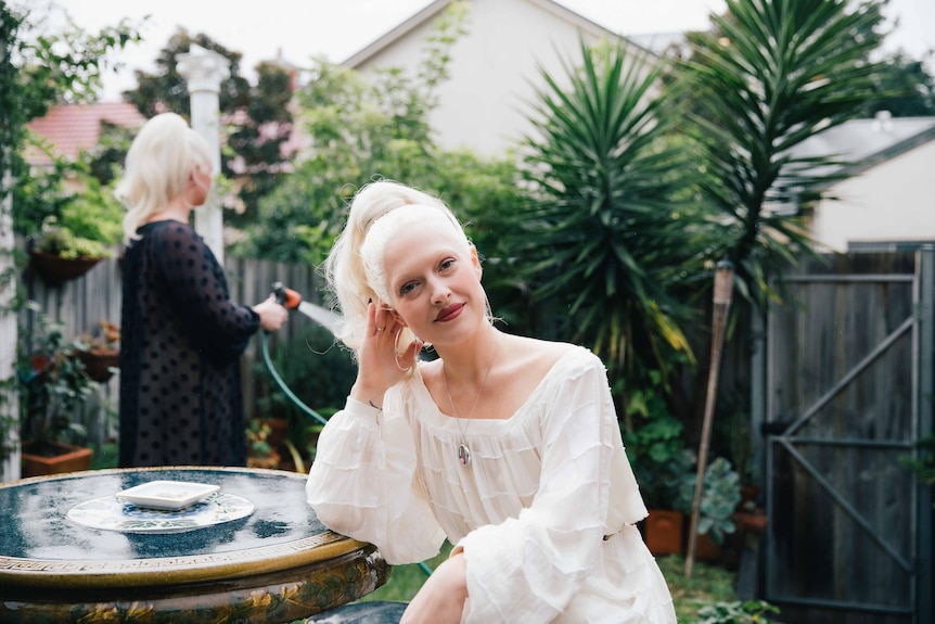 A glamorous white woman with platinum blonde hair, wearing a sheer white dress sits at a garden table.