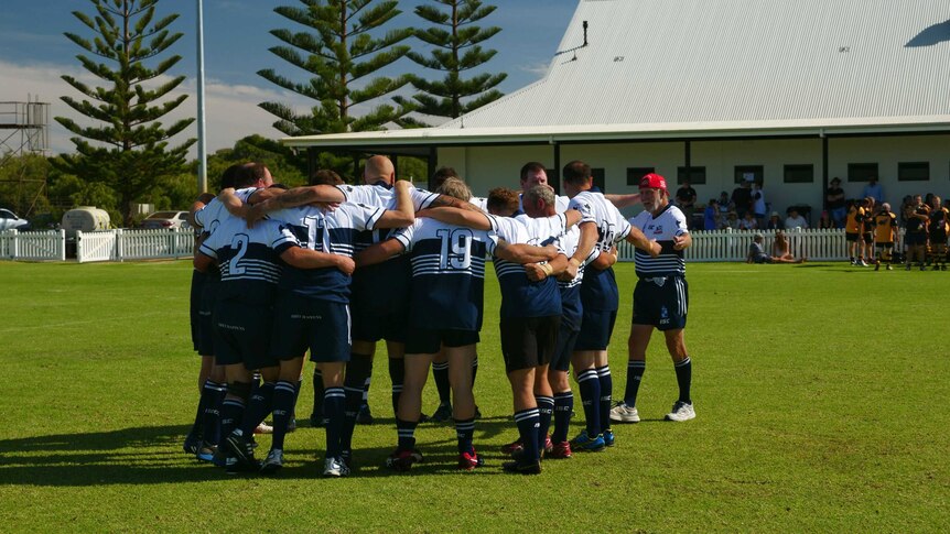 A group of rugby players in a circle with an older man coming to join the team circle.