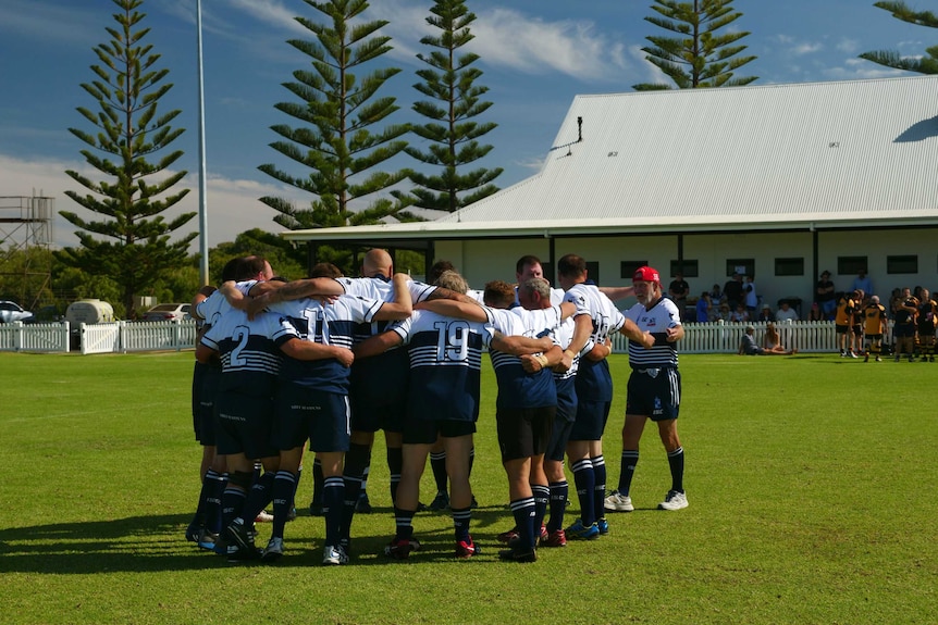 A group of rugby players in a circle with an older man coming to join the team circle.