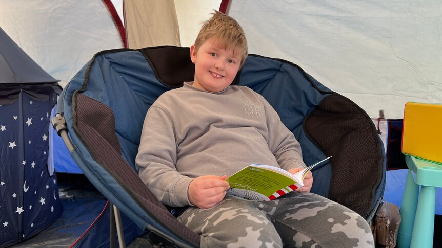 A smiling boy sits on a large chair in a tent, reading a Dr Seuss book.
