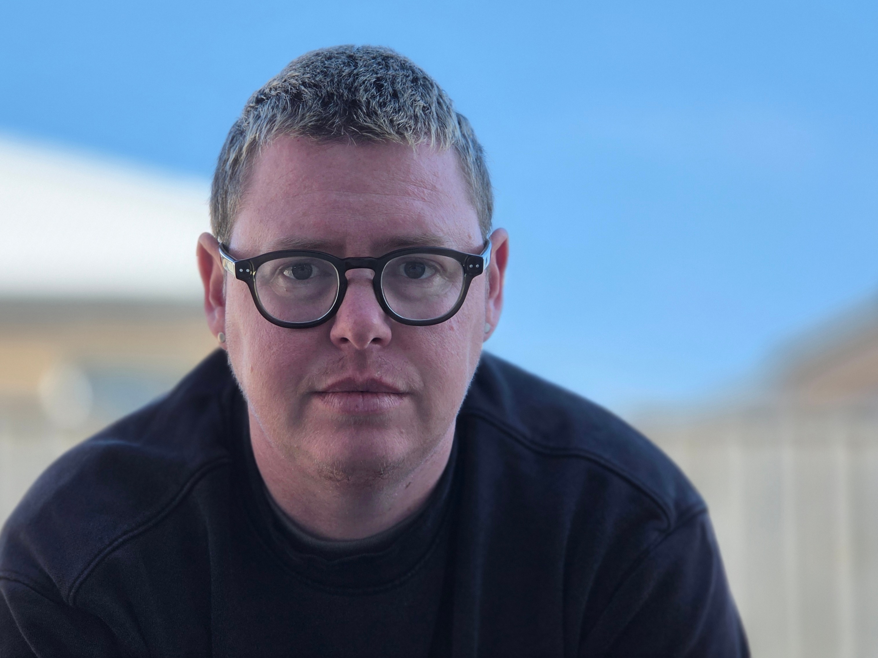 A person wearing a black shirt and black glasses looks seriously at the camera.