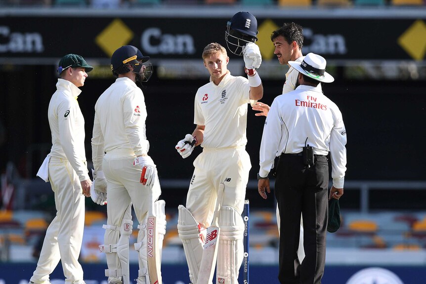 Joe Root holds his broken helmet in the air, surrounded by players and umpires.