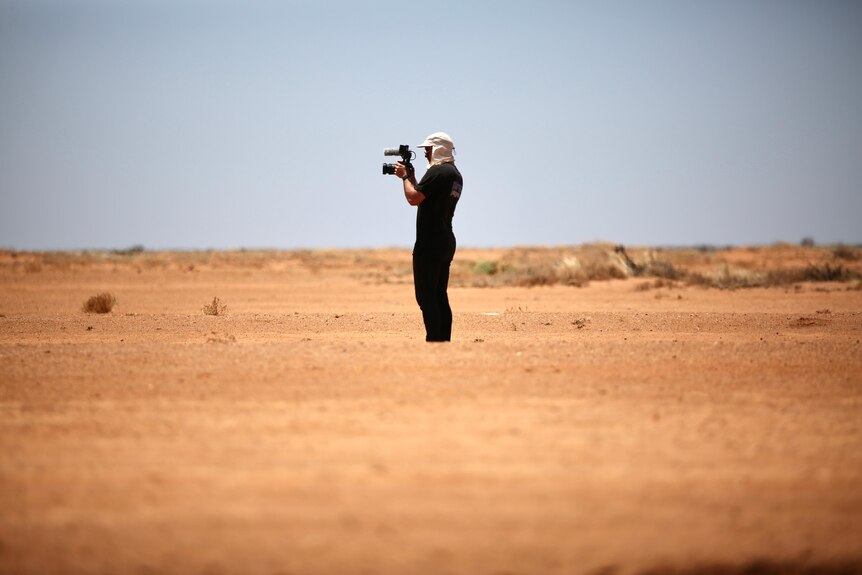 A man wearing a white flap cap and black t-shirt and pants stands in the distance with a camera in a desert setting.