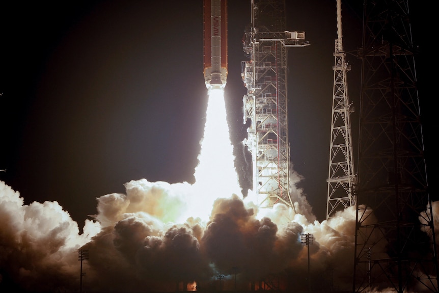 A US moon rocket blasts off vertically from a launncher system against a dark sky.