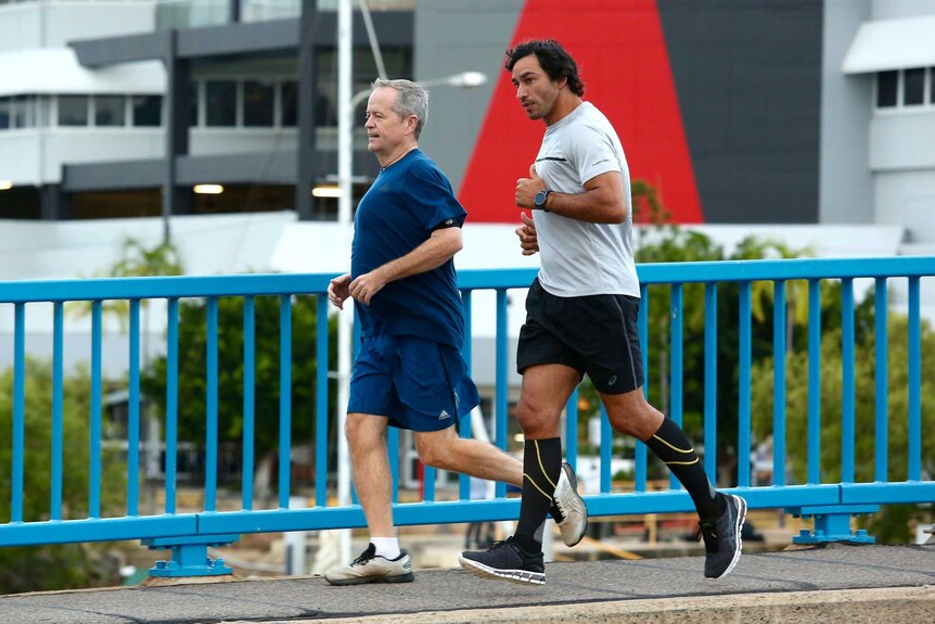 The men run side by side along a bridge with a blue fence