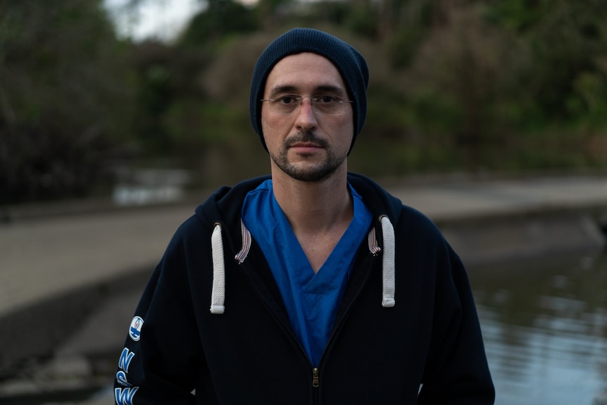 Male nurse wearing a beanie and hoodie over his scrubs, standing outside.