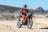 Sand flies up as a motorcyclist speeds through rocky terrain on the first leg of a rally race.