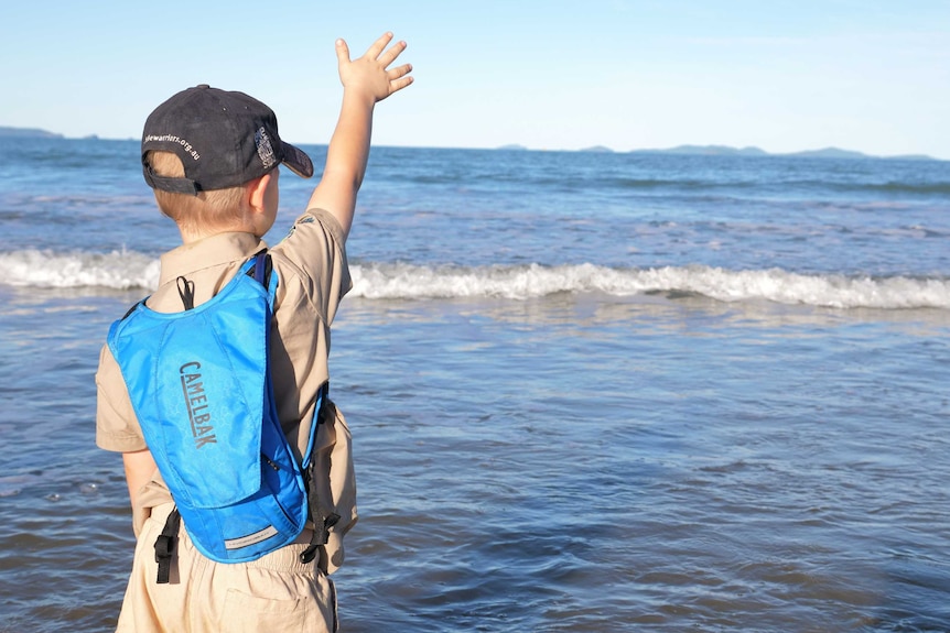Owen with his khaki wildlife warrior uniform, a blue backpack and a dark cap on waves at the ocean.