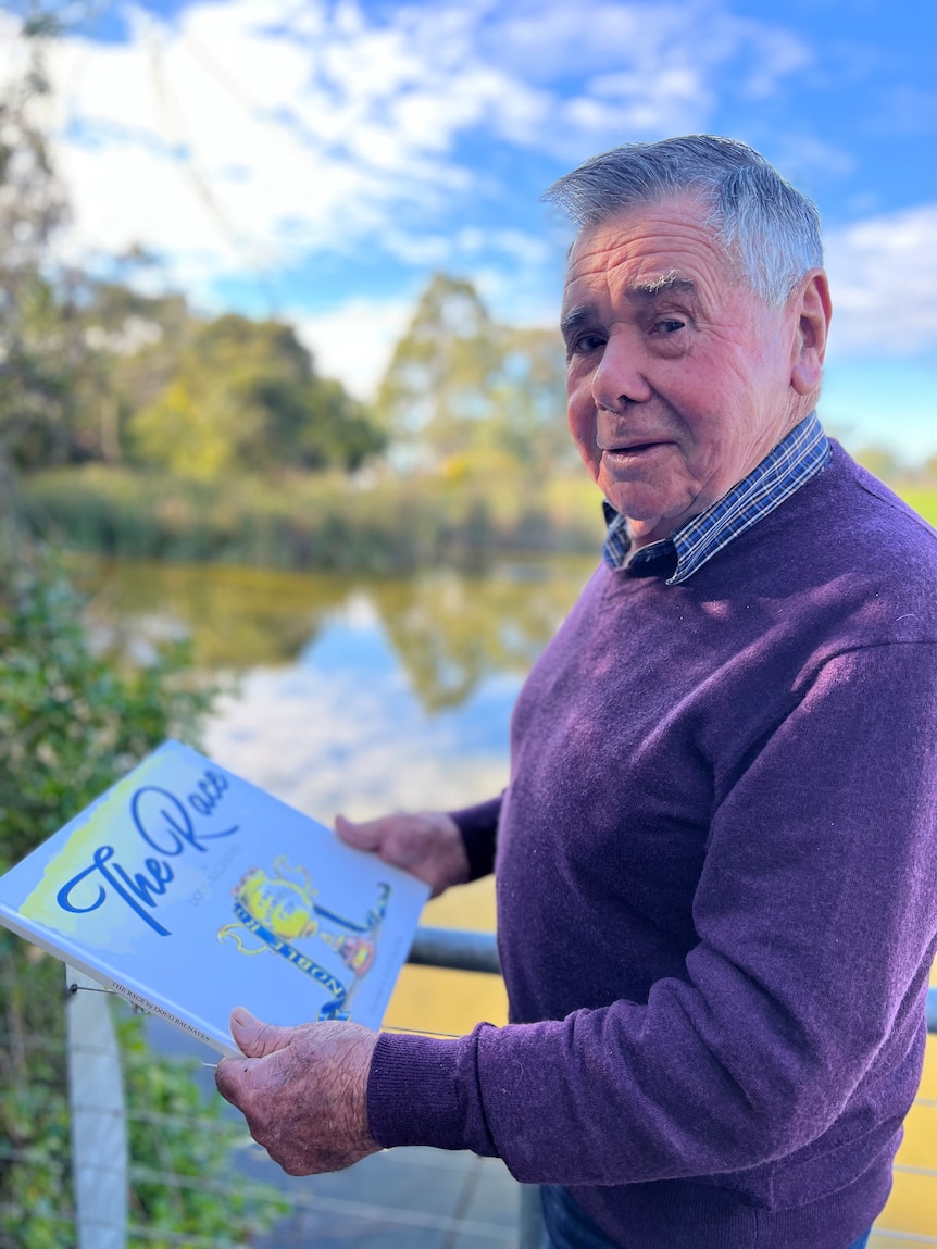 An elderly man in a purple jumper holds a book titled The Race in a rural setting