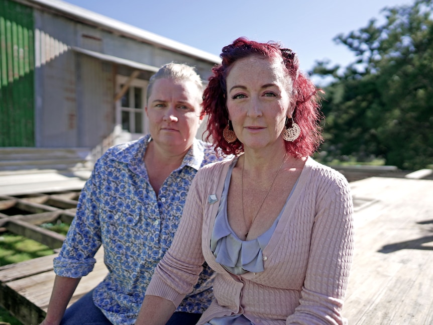 Two women sit on an unfinished deck outside a converted shed. They are looking at the camera with neutral expression.