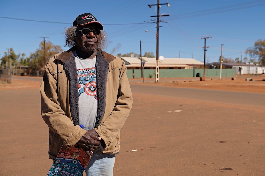 Warlpiri elder Warren Japanangka Williams wearing a jacket, hat and sunglasses looks across the camera.