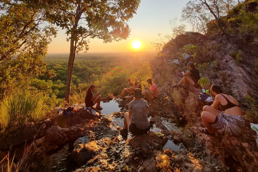 Women in hiking and swimming gear sit on rocks looking out at sunset.
