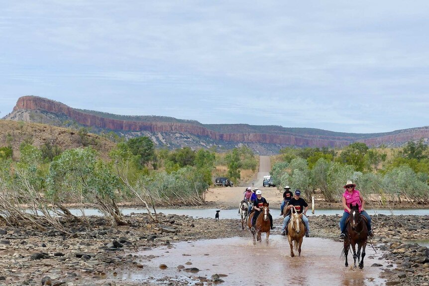 Group of nine horse riders wading through the Pentecost River with the Cockburn Range in the background.