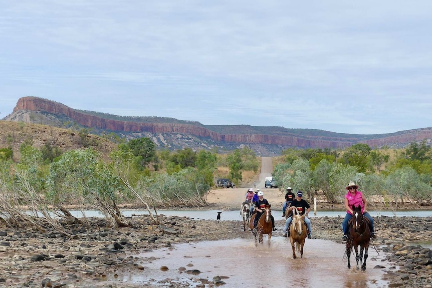 Group of nine horse riders wading through the Pentecost River with the Cockburn Range in the background.