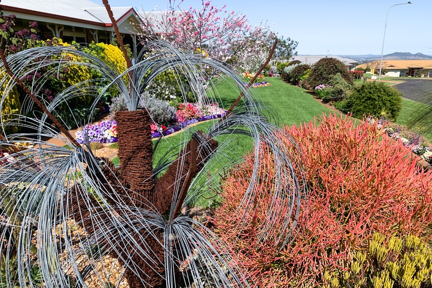 a metal grass tree sculpture in a garden bed surrounded by shrubs and flowers