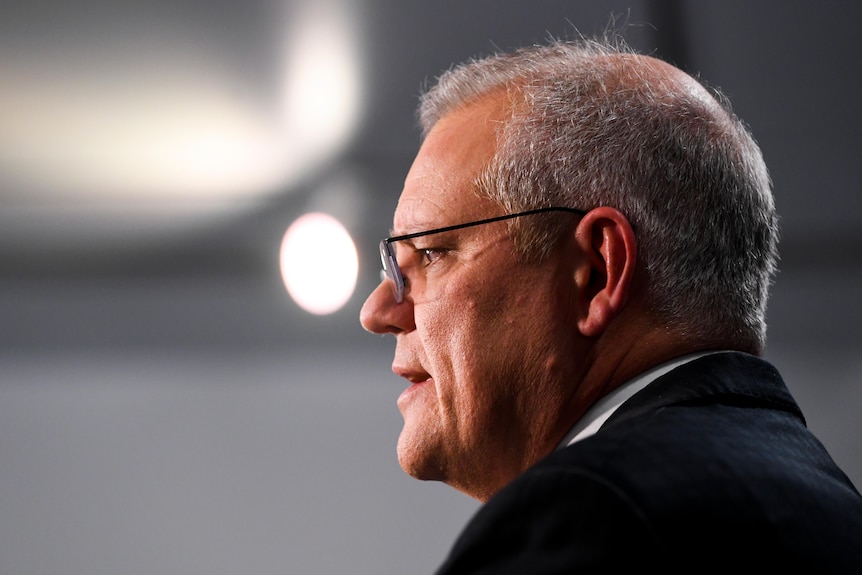 A man in a suit wearing glasses stands side-on while talking.