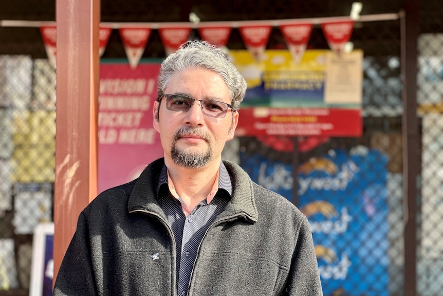 A bespectacled man with grey hair and a neat beard standing in front of a shop in a country town.