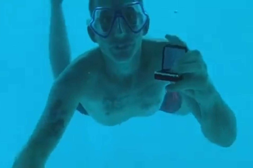A man wearing a diving mask shows an open ring box to the camera while underwater.