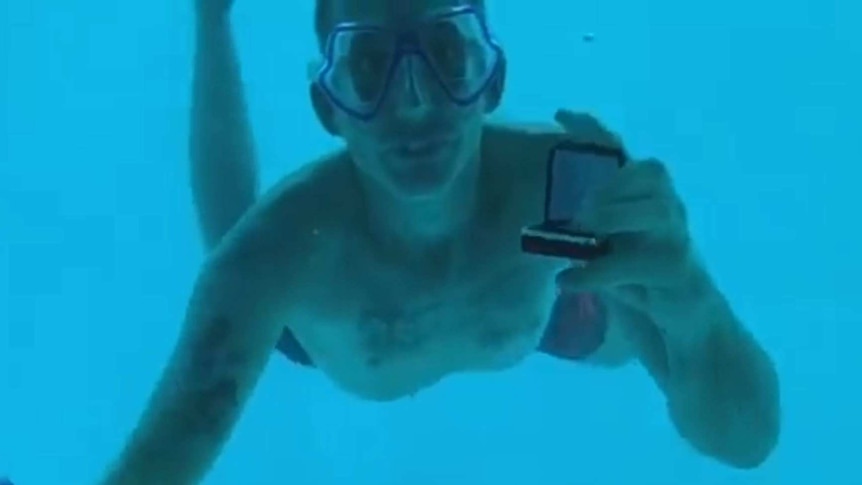 A man wearing a diving mask shows an open ring box to the camera while underwater.