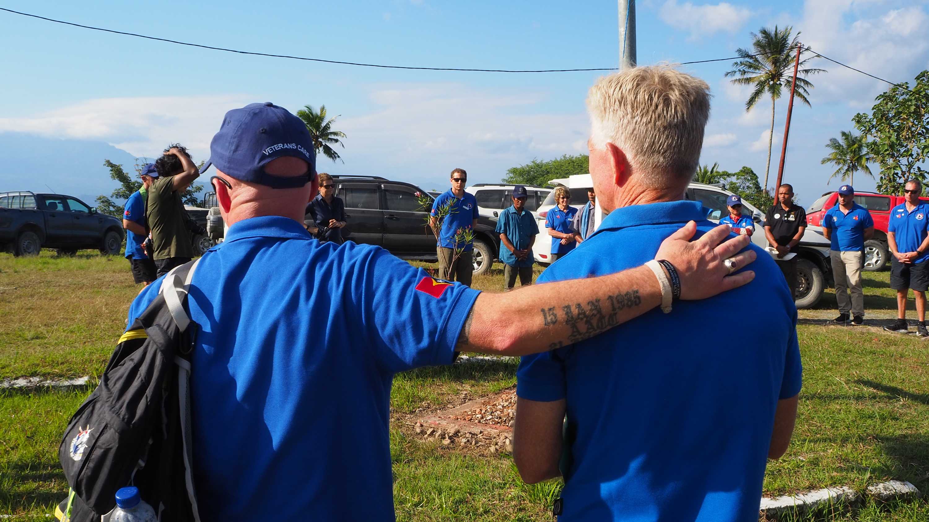 Two men in the same blue shirts stand side by side during a ceremony, one has his hand on the others back