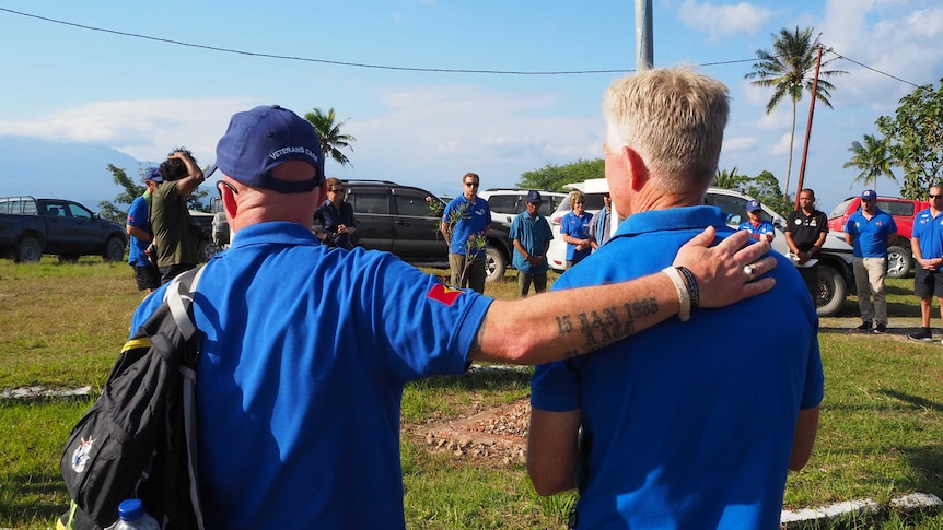 Two men in the same blue shirts stand side by side during a ceremony, one has his hand on the others back