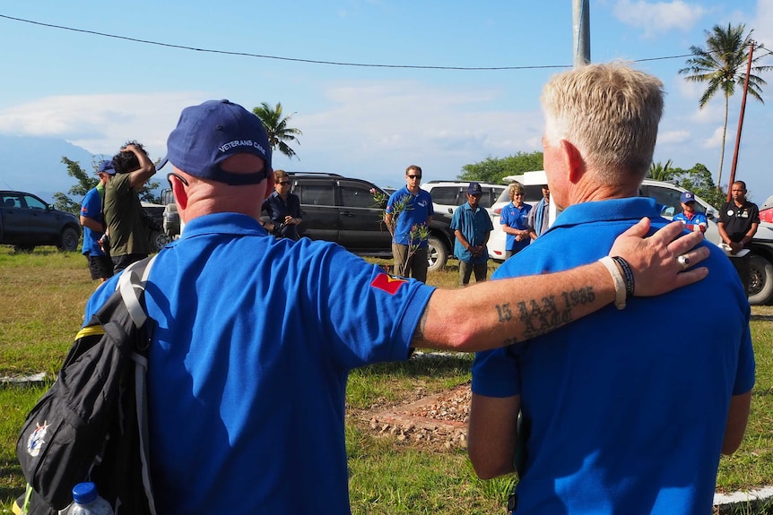 Two men in the same blue shirts stand side by side during a ceremony, one has his hand on the others back