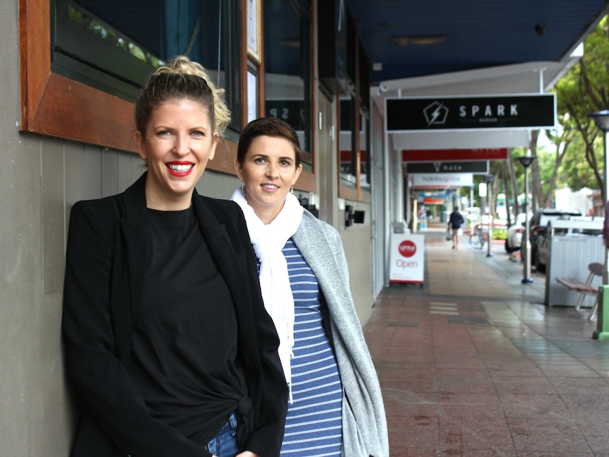 Two sisters stand smiling at the top of a street with lots of businesses and people walking in the background.