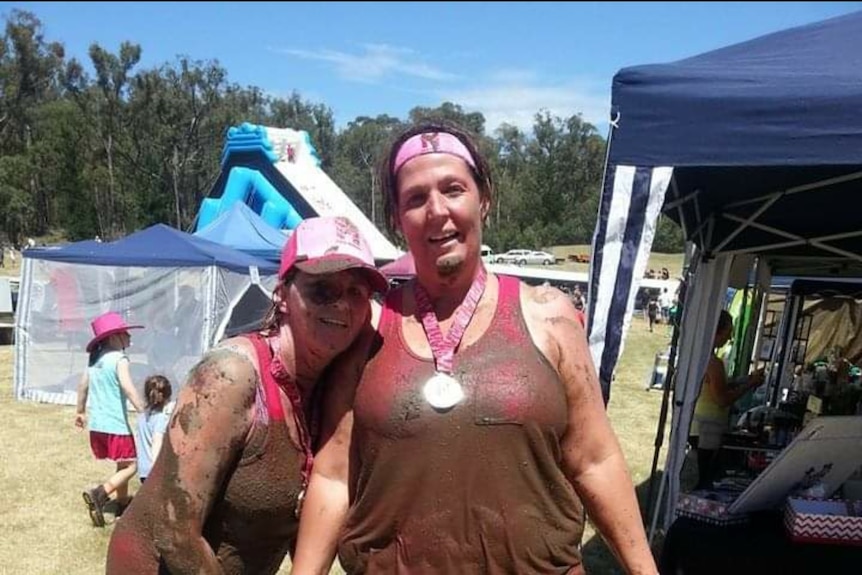 Two women dressed in pink and covered in mud smiling at the camera with tents in the background.