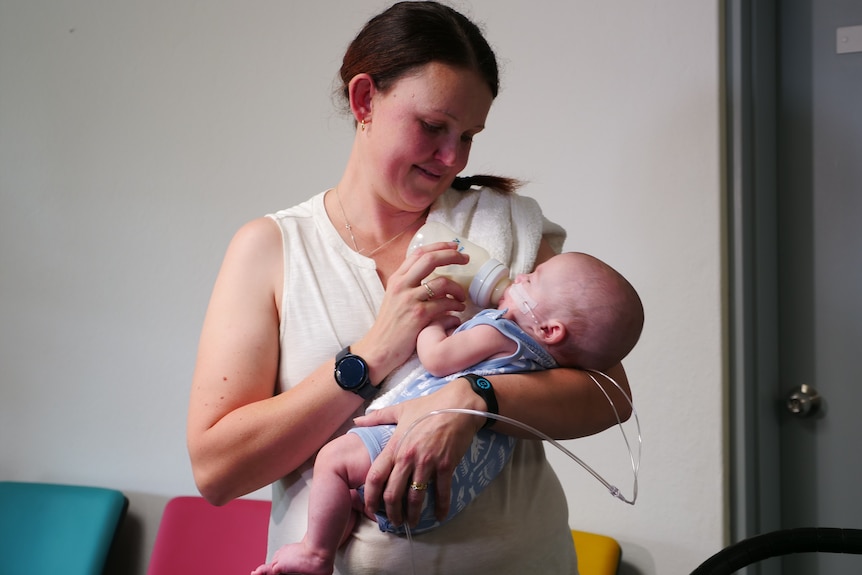 A woman with long brown hair holds a baby boy and feeds him a bottle.