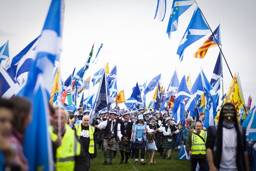 Hundreds of protesters march across a lawn in kilts, masks and waving blue flags. Some play bagpipes.