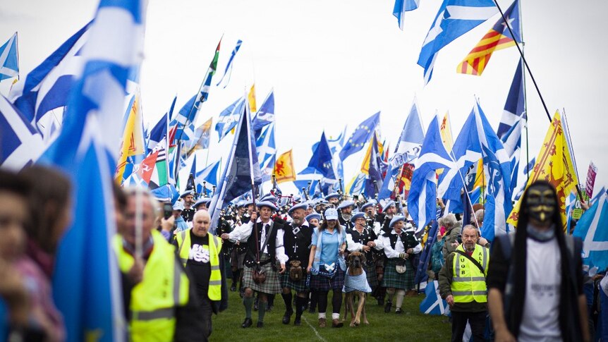 Hundreds of protesters march across a lawn in kilts, masks and waving blue flags. Some play bagpipes.