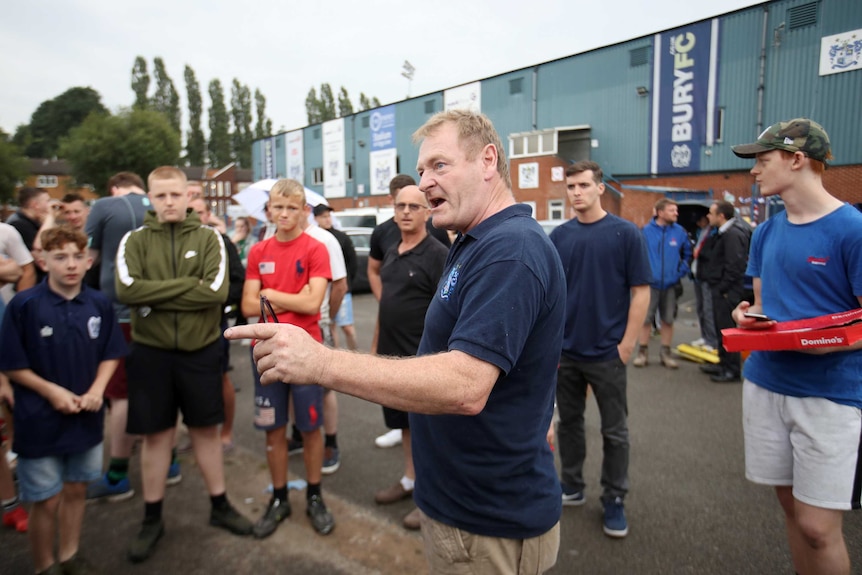 An angry fan points and shouts as other fans stand around him with arms folded outside Bury FC's stadium