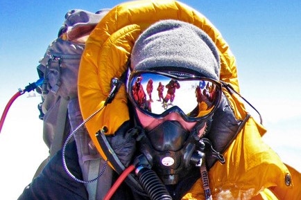 A man wearing a jacket and layers of thermal clothing, a beanie, oxygen mask and goggles at the top of a mountain