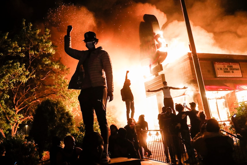 Protesters raise their fists in defiance outside a burning fast-food restaurant.