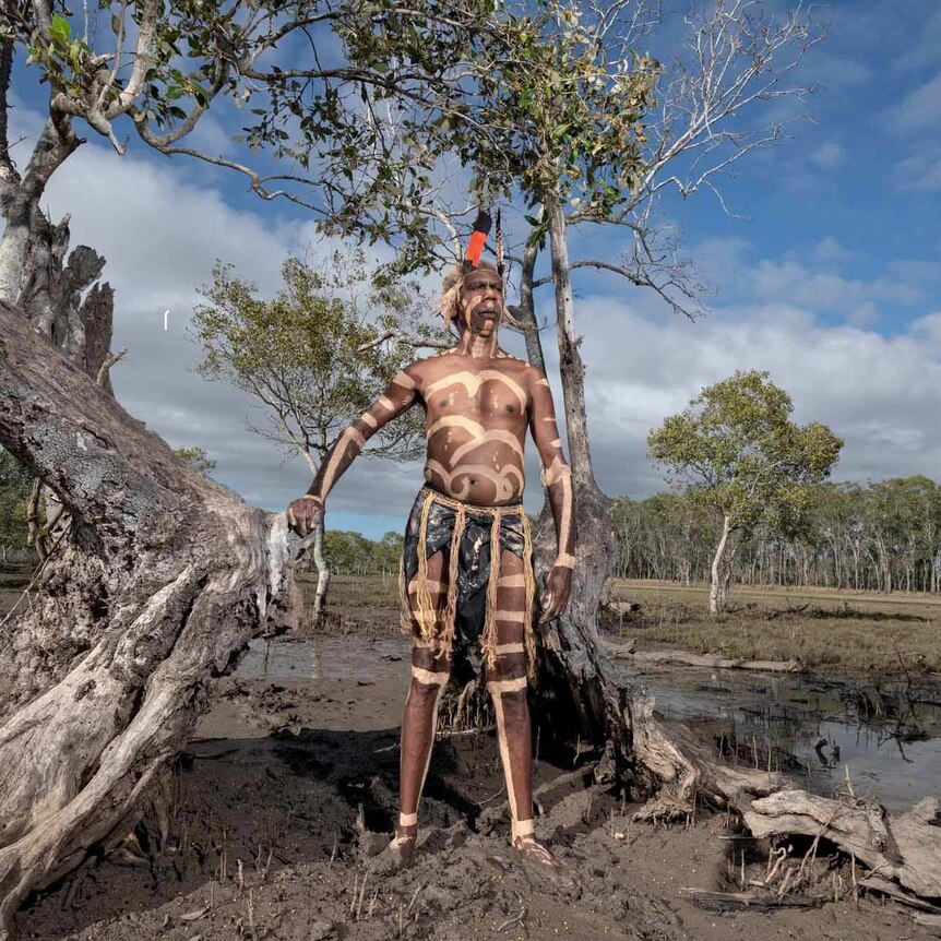 An Aboriginal man, painted with ochre, standing in front of a mangrove tree.