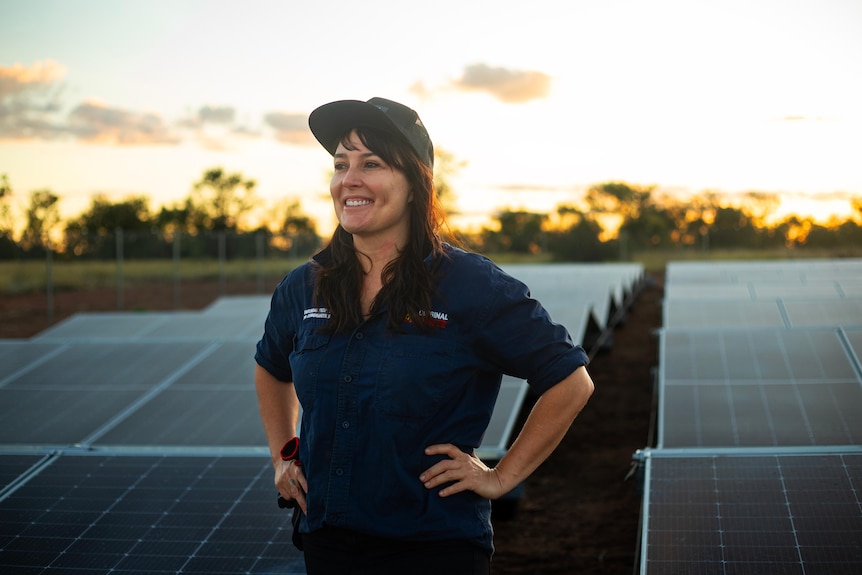 A photo showing a  woman standing with hands on hips in middle of field containing solar panel grid