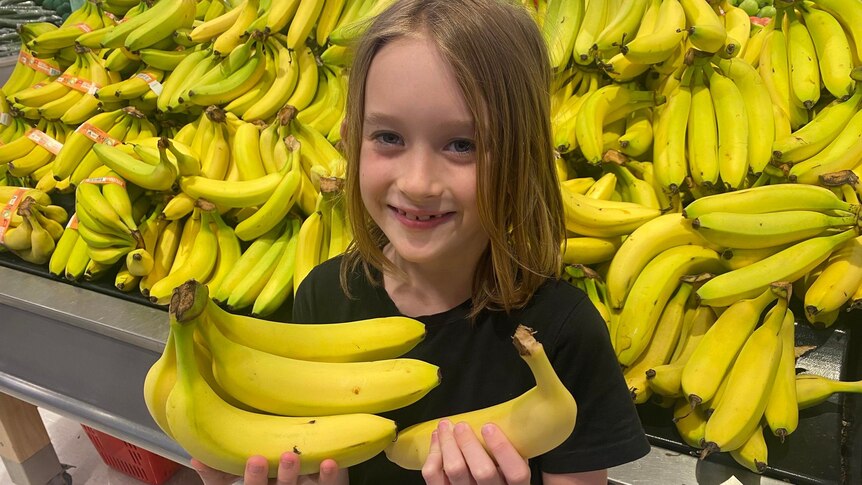 A young boy with shoulder-length hair stands in front of bananas at the supermarket holding bananas in both hands