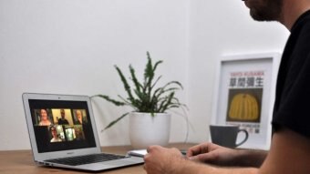 A man sits at a table participating in a Zoom meeting. On the laptop in front of him are five faces of people in the meeting.