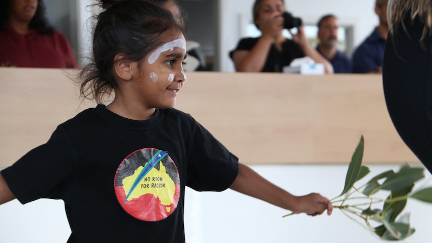 A young gunditjmara girl in t-shirt that has map of aus saying no room for racism, dancing with gum leaves