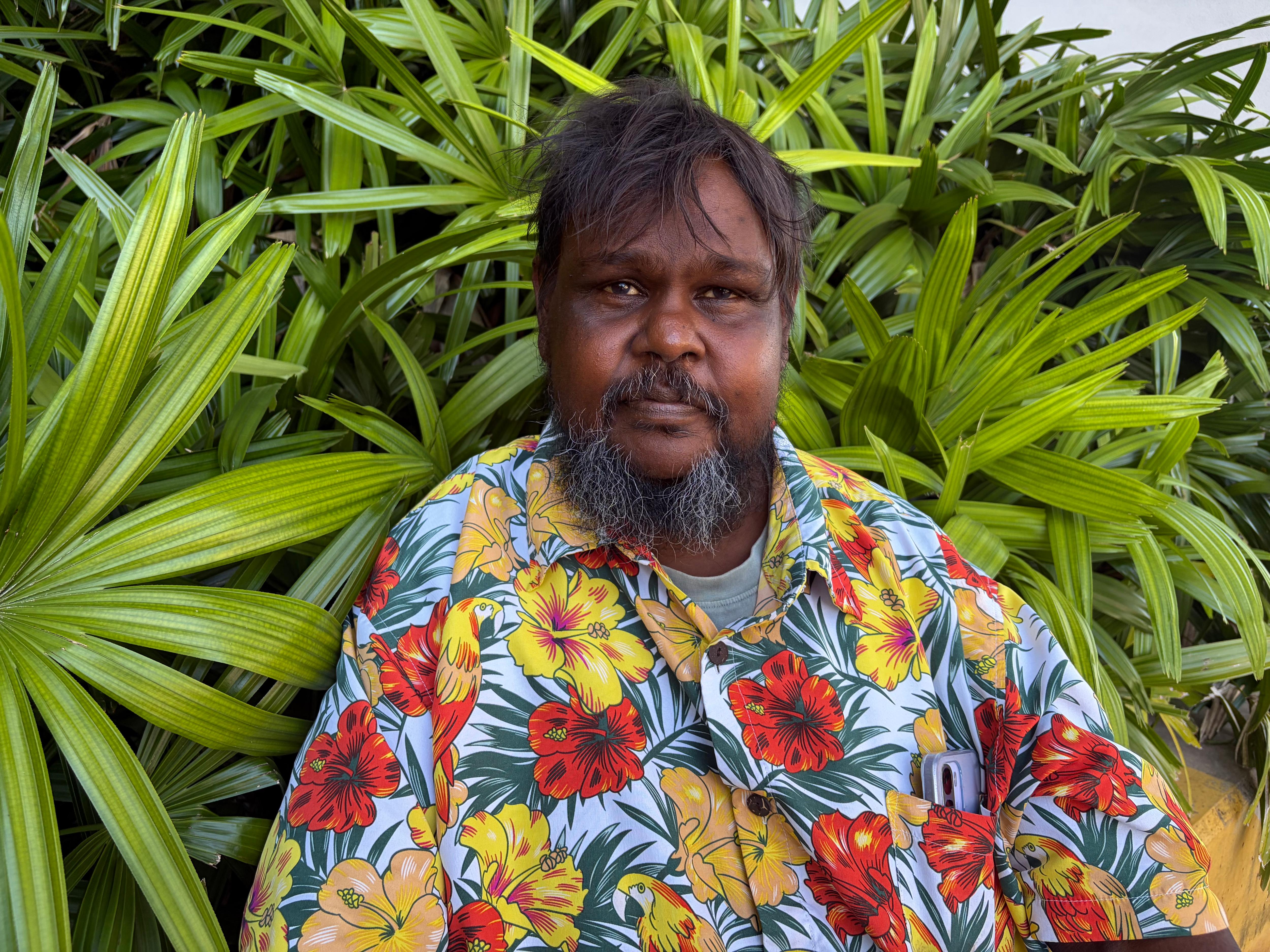 Aboriginal man, short brown hair with fringe over forehead, wearing Hawaiian shirt, sitting among green leaves.