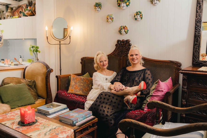 Two white women with platinum blonde hair and wearing semi-sheer dresses sit on a velvet couch in a lavishly decorated room.